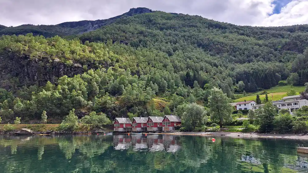Red boathouses reflected in still green water below a wooded slope